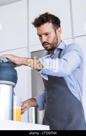 Dal basso dello chef adulto maschio in grembiule guardando in basso mentre si è seduti al tavolo della cucina con spremiagrumi durante il sano processo di colazione alla luce del giorno Foto Stock