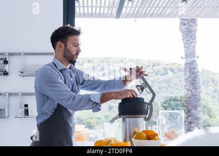 Vista laterale dello chef adulto adulto in grembiule che guarda in basso mentre si è seduti al tavolo della cucina e si mette l'arancia nello spremiagrumi durante una pausa salutare Foto Stock