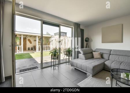 Divano grigio vuoto e confortevole collocato da una grande e luminosa finestra che si affaccia sul cortile sul retro del soggiorno di una casa moderna Foto Stock
