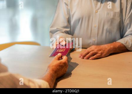 Tagliate le mani di un anziano anonimo in camicia bianca che riceve il passaporto dall'agente di registrazione durante il check-in per il volo all'aeroporto Foto Stock