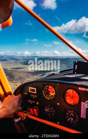 La mano di un pilota viene vista sui comandi di un piccolo aereo, volando sopra un paesaggio mozzafiato in una giornata nuvolosa Foto Stock