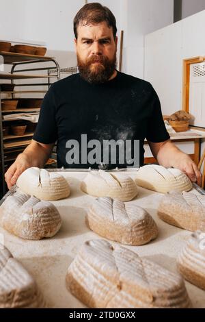 Ritratto di un fornaio maschio maturo vestito con casuali neri che tengono in mano un vassoio da forno con impasti di pane crudo mentre si trova in cucina alla casa di produzione Foto Stock