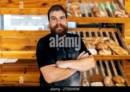 Ritratto di un uomo maturo con barba sicura di sé con le braccia incrociate contro gli scaffali di legno con pane appena sfornato nel forno Foto Stock