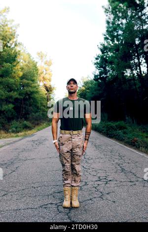Tutta la lunghezza di un commando di forza speciale sicuro di mezza età che indossa pantaloni mimetici e t-shirt verde in posizione di attenzione sulla strada nella foresta Foto Stock