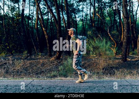 Vista laterale completa del corpo di un commando di mezza età che indossa uniforme e zaino che corre lungo la strada contro gli alberi della foresta Foto Stock