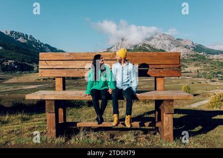 Una coppia di hipster sorridenti siede da vicino su una grande panchina rustica in legno, tenendosi per mano e guardandosi con un pittoresco paesaggio montano in t Foto Stock