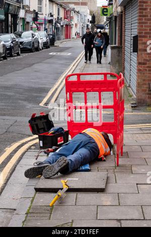 Un ingegnere BT Openreach sdraiato su un marciapiede che lavora su cavi telefonici sotterranei a cui si accede sollevando una copertura di ispezione su una strada Foto Stock