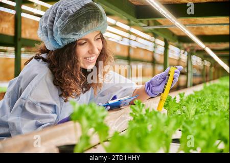 Donna scienziata che usa un pHmetro in serra. Gioiosa giardiniera che tiene un portaappunti e sorride misurando il valore del pH del terreno con un dispositivo elettronico digitale. Foto Stock