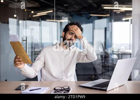 Ritratto di un giovane indiano che lavora in ufficio con un portatile, tiene la testa con la mano, guarda la fotocamera sotto shock, riceve cattive notizie per lettera. Foto Stock