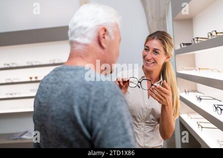 Un'ottica che mostra gli occhiali a un cliente senior di sesso maschile. concentrati sulla donna con occhiali in un negozio ottico pieno di scaffali. Foto Stock