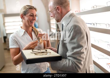 Donna sorridente che esamina gli occhiali con un ottico in un negozio ottico. Ripiani per occhiali sullo sfondo. Foto Stock