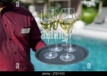 Cameriere con una camicia rossa che tiene in mano un vassoio con tre bicchieri di champagne a bordo piscina. Immagine del concetto di viaggio alberghiero Foto Stock