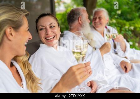Un gruppo di persone con accappatoi bianchi che ridono e tengono in mano dei bicchieri da champagne per festeggiare su un lettino in un hotel spa e benessere Foto Stock