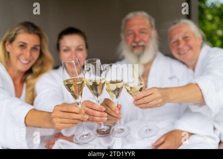 quattro persone in accappatoi bianchi, brindate con bicchieri da champagne e festeggiate una giornata di benessere in hotel Foto Stock