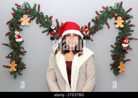 Donna latina con maschera protettiva, maglione e cappello di natale su sfondo grigio con decorazione natalizia, nuovo normale Foto Stock