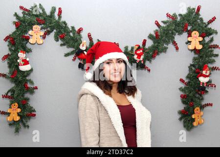 Donna latina con maschera protettiva, maglione e cappello di natale su sfondo grigio con decorazione natalizia, nuovo normale Foto Stock