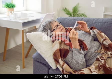 Donna anziana malata che ha freddo e febbre prende la sua temperatura mentre è sdraiata sul divano Foto Stock