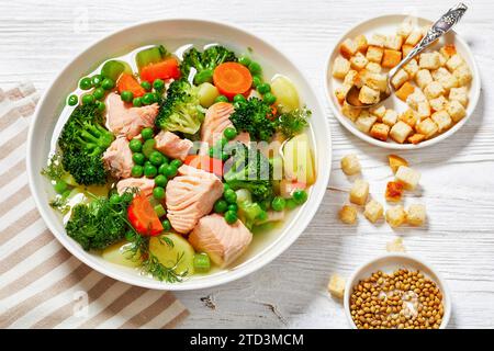 salmone, piselli, broccoli, patate, sedano, zuppa di carote ed erbe verdi in ciotola bianca su tavolo di legno bianco, vista orizzontale dall'alto Foto Stock