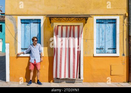 Giovane uomo appoggiato contro il muro della casa, facciata gialla della casa con porta d'ingresso con tenda rossa e bianca e finestre con persiane blu, colorate Foto Stock