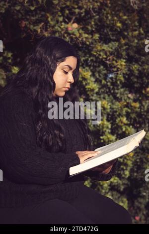 Giovane donna, seduta attenta a leggere un libro in giardino, foto con vista laterale a mezzo corpo verticale Foto Stock