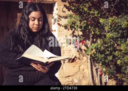 Giovane ragazza latina seduta a leggere un libro nel giardino vicino alla casa di campagna Foto Stock