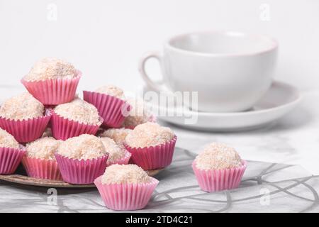 Palline di rum di cocco in stampi rosa su un piatto accanto a una tazza di caffè vuota su una superficie marmorizzata Foto Stock