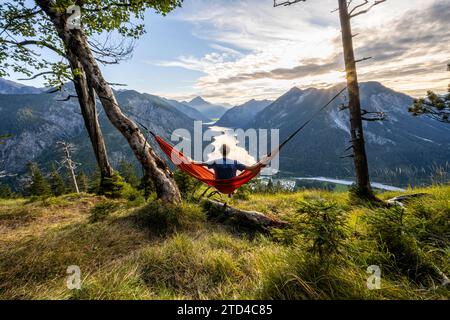 Giovane seduto su un'amaca arancione, vista di Plansee da Schoenjoechl, al tramonto con stella del sole, vista panoramica delle montagne con il lago, Plansee Foto Stock