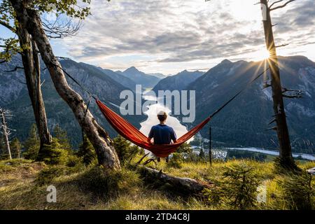 Giovane seduto su un'amaca arancione, vista di Plansee da Schoenjoechl, al tramonto con stella del sole, vista panoramica delle montagne con il lago, Plansee Foto Stock