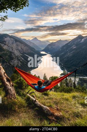 Giovane seduto su un'amaca arancione, vista di Plansee da Schoenjoechl, al tramonto, vista panoramica delle montagne con il lago, Plansee, Tirolo, Austria Foto Stock