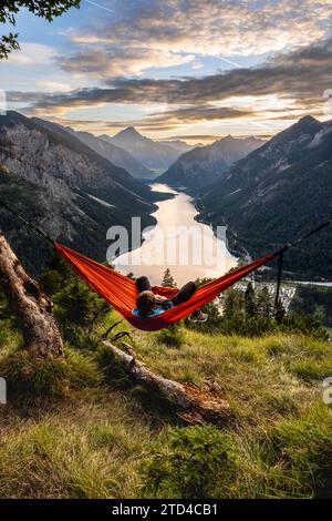 Giovane seduto su un'amaca arancione, vista di Plansee da Schoenjoechl, al tramonto, vista panoramica delle montagne con il lago, Plansee, Tirolo, Austria Foto Stock