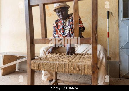 Lavoro di primo piano di tessitura di una sedia di legno da parte di un uomo africano cieco con cappello sulla testa. Foto Stock