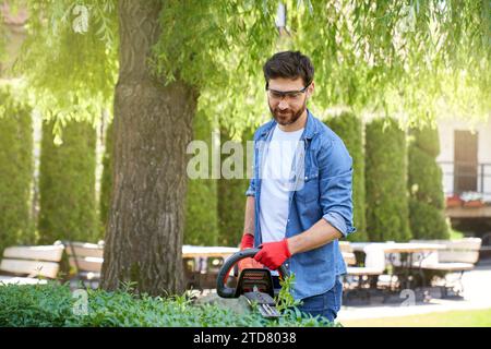 Giardiniere maschio concentrato nei guanti per tagliare siepi in bosso con il rifinitore sul cortile. Vista frontale dell'uomo sorridente colletto blu che utilizza attrezzature moderne, mentre la vista è Foto Stock