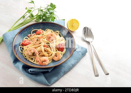 Spaghetti con gamberetti e pomodori in salsa di limone con contorno di prezzemolo in un piatto blu su un tavolo di legno bianco, pasto mediterraneo a base di pesce fatto in casa, Foto Stock