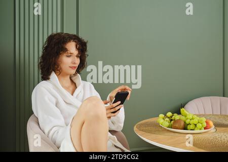 Una giovane donna che compone il numero della sua amica mentre è seduta al tavolo dopo la doccia Foto Stock