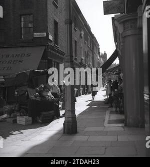 Anni '1950, storica, una donna che fa shopping con un commerciante di sesso maschile al Market Stall, Shepherd Market, a Mayfair, Londra, Inghilterra, REGNO UNITO. All'angolo del vicolo lastricato, Hayward Stores e un cartello per una buona casa magazzino a 1'9 al mese. Situato a Mayfair, in prossimità di Piccadilly, Shepherd Market è una parte affascinante del centro di Londra con la sua piccola piazza e le sue strette stradine laterali, essendo conosciuto come un "villaggio a Piccadilly". Foto Stock