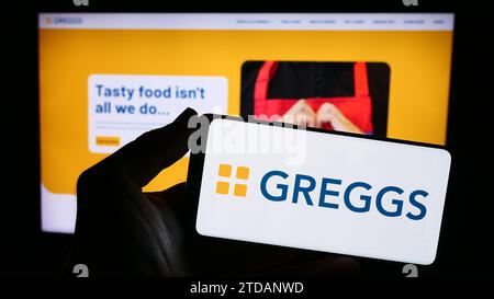 Persona che possiede uno smartphone con il logo della società britannica di panetteria Greggs plc davanti al sito Web. Concentrarsi sul display del telefono. Foto Stock