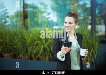 lavoratrice moderna sorridente vicino al business center in una giacca nera con una tazza di caffè utilizzando lo smartphone. Foto Stock