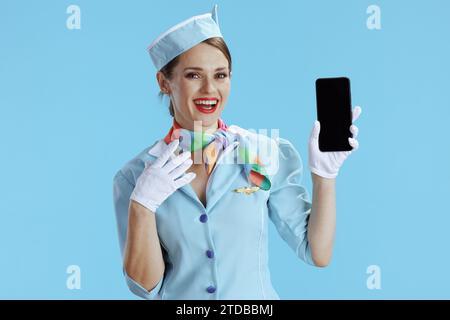 donna sorridente ed elegante hostess isolata su sfondo blu in uniforme blu che mostra lo schermo vuoto dello smartphone. Foto Stock