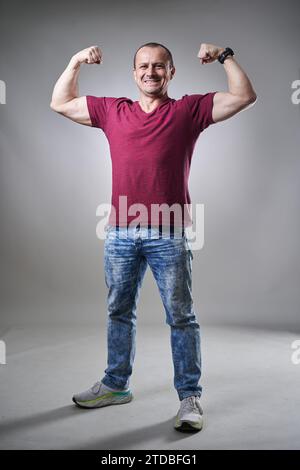 Uomo di mezza età con t-shirt e jeans che piegano i bicipiti su sfondo grigio Foto Stock