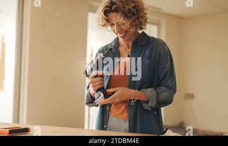 La donna sorride mentre tiene in mano una pistola perforatrice in una cucina rinnovata. Impegnata in un progetto di miglioramento della casa fai-da-te, aggiorna con sicurezza la sua casa e i suoi spettacoli Foto Stock