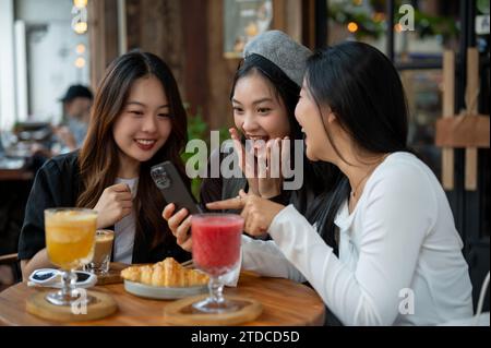 Un gruppo di attraenti amiche asiatiche è sorpreso da alcune notizie online su uno smartphone e si diverte a parlare in un bar o in un ristorante, a uscire Foto Stock