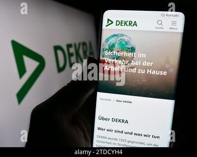 Persona che possiede il cellulare con la pagina Web dell'organizzazione tedesca di esperti DEKRA davanti al logo aziendale. Concentrarsi sul centro del display del telefono. Foto Stock