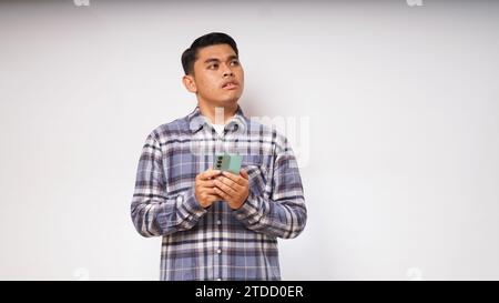 Giovane asiatico che tiene il suo telefono cellulare con un'espressione triste e confusa su sfondo bianco. foto studio Foto Stock