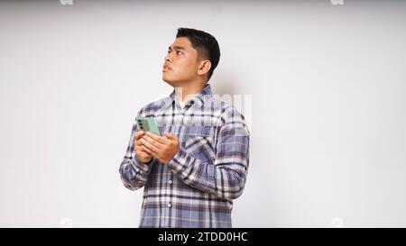 Giovane asiatico che tiene il suo telefono cellulare con un'espressione triste e confusa su sfondo bianco. foto studio Foto Stock