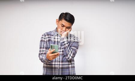 Giovane asiatico che tiene il suo telefono cellulare con un'espressione triste e confusa su sfondo bianco. foto studio Foto Stock