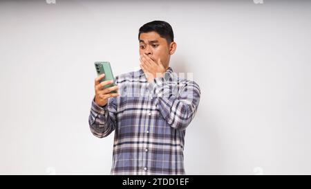 Giovane uomo asiatico che mostra l'espressione del viso scioccata mentre si utilizza il telefono cellulare su sfondo bianco Foto Stock