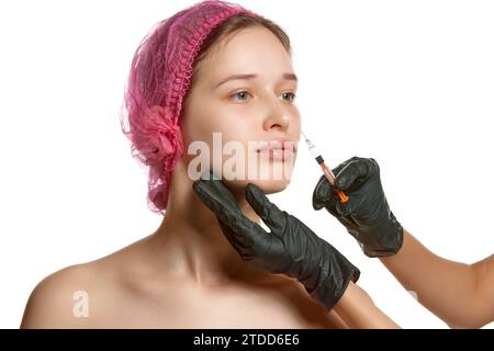 Ritratto di una bella donna in berretto rosa per trattamenti cosmetici che fanno iniezioni di bellezza. Le mani del medico con guanti neri tengono la siringa. Lifting facciale. Foto Stock