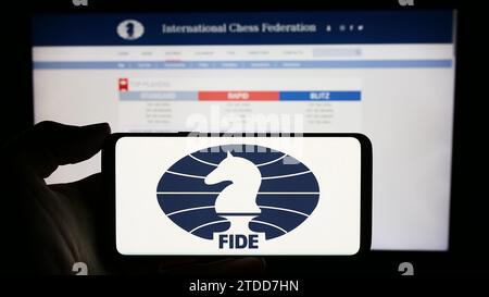 Persona in possesso di un cellulare con il logo dell'organizzazione International Chess Federation (FIDE) davanti alla pagina web. Concentrarsi sul display del telefono. Foto Stock