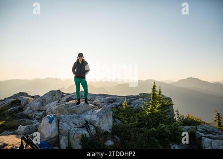 donna felice e in forma in piedi sulla cima rocciosa della montagna al tramonto Foto Stock