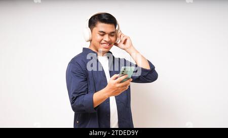 Un giovane asiatico felice che indossa cuffie wireless che ascolta musica dallo studio dello smartphone scattato isolato su sfondo bianco Foto Stock